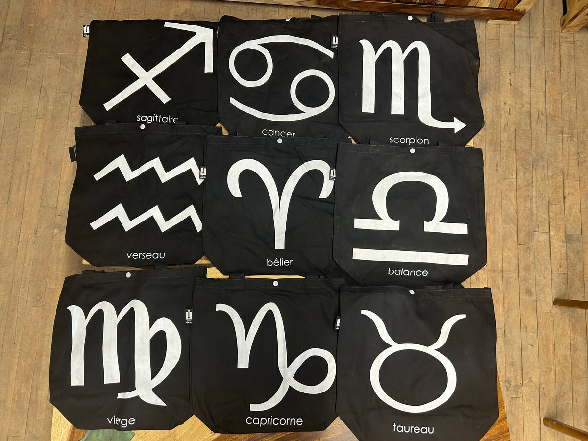 Neuf tote bags noirs imprimés de signes du zodiaque, disposés au sol